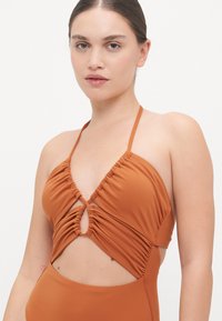 Maillot de bain couleur rouille avec un design froncé, des bonnets triangulaires et une découpe centrale, doté de fines bretelles et d'une texture lisse et extensible.
