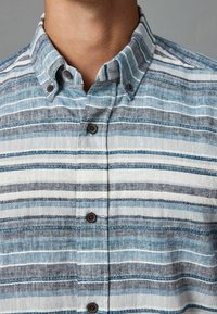 Koszula na guziki z paskowanym wzorem w odcieniach niebieskiego i szarości, z teksturowanym materiałem oraz brązowymi guzikami przy kołnierzyku i patce przy guzikach.