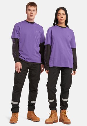 T-shirt viola a maniche corte sovrapposto a camicie a maniche lunghe nere, con pantaloni neri e stivali marroni. Accenti riflettenti sui pantaloni.