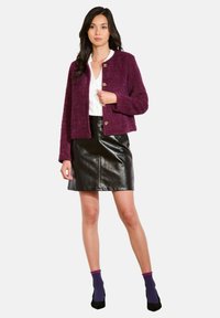 Veste courte en velours violet avec un design court, dotée de boutons dorés, associée à une jupe en cuir noir et des chaussettes à motifs.