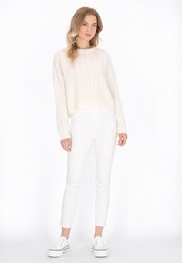 Maglione a treccia di colore crema, lunghezza cropped, abbinato a jeans skinny bianchi e sneakers bianche con plateau. Design semplice e pulito con una texture morbida.