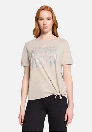 Ung kvinde med rødt hår iført beige top med korte ærmer, sølvfarvet bølgemønster og knude ved kanten, kombineret med sorte bukser.