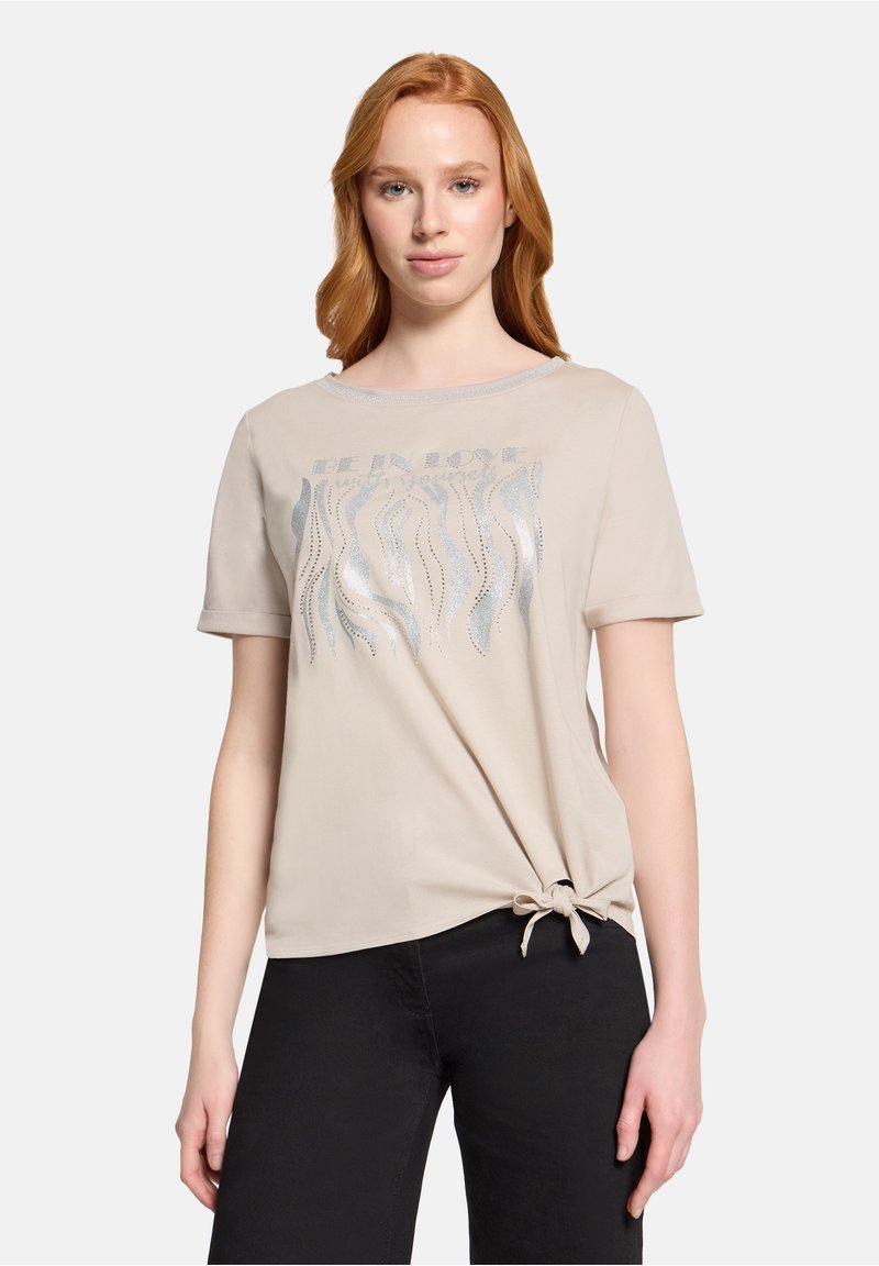 Jeune femme aux cheveux rouges portant un haut beige à manches courtes avec un motif argenté en forme de vague et un nœud noué à l'ourlet, associée à un pantalon noir.