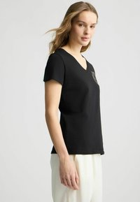 Zwarte V-hals T-shirt met korte mouwen, gemaakt van zachte stof. Heeft een klein logo op de borst. Model staat zijwaarts, draagt lichte broek.