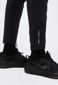 Πόδια που φορούν μαύρο παντελόνι Therma-Fit και μαύρα αθλητικά παπούτσια Nike με πλέγμα και ταινίες έλξης σε λευκό φόντο.