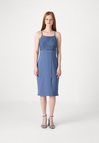 Zila kleita ar mežģīņu augšu, piegulošu vidukli un ceļa garuma svārkus ar sānu šķēlumu. Modelis valkā atbilstošas zilas kurpes ar papēžiem.
