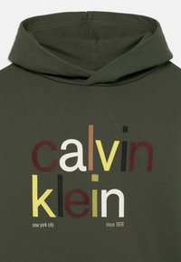 Olivazöld kapucnis pulóver elől „calvin klein” felirattal, különböző színekben és betűtípusokkal, beleértve a fehéret, sárgát, barnát és feketét.