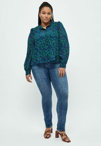 Blusa floreale multicolore in blu navy e verde con maniche a palloncino, abbinata a jeans blu aderenti e sandali con tacco marroni.