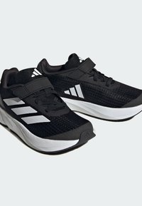 Zapatillas deportivas negras con una parte superior texturizada, tres rayas blancas en el costado y una suela blanca. Cuenta con una correa de velcro y un sistema de cordones.