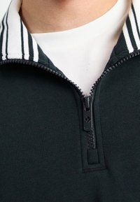 Donkergroene ritsjack met een gestreepte kraag, uitgerust met een zwarte rits en een zachte, textuur. De rits loopt iets onder de kraag door.