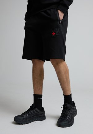 Mies, jolla mustat shortsit, joissa Kanadan logo, mustat Nike-sukat ja mustat Nike-tossut, seisoo yksinkertaisen vaalean taustan edessä.