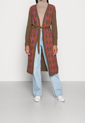 Personne portant un long cardigan marron avec des motifs losanges rouges et violets, un col roulé beige, un jean bleu clair et des baskets blanches.
