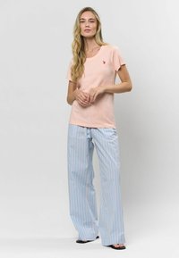 Svetloružové tričko s krátkym rukávom a malým červeným logom na hrudi, spárované s modro-bielymi širokými nohavicami s vertikálnym pruhom.