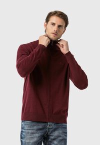 Burgunderfarbener Reißverschluss-Pullover mit hohem Kragen, taillierter Passform, gerippten Bündchen und Saum, aus einem weichen, strukturierten Strickmaterial.