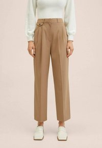 Pantalon marron sur mesure avec taille haute, plis et bouton décoratif. Tissu lisse et coupe droite. Porté avec un haut blanc et des chaussures.