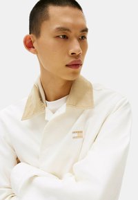 Giacca bianca con colletto in corduroy beige, dotata di una piccola toppa con logo. Tessuto morbido e vestibilità rilassata con chiusura abbottonata.