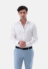 Weißes Langarmhemd mit klassischem Kragen, durchgehender Knopfleiste und glatter Textur, kombiniert mit hellblauen Hosen und einem schwarzen Gürtel.