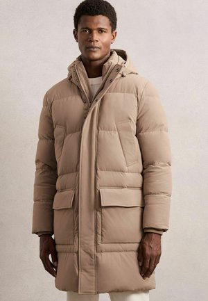 Mees, kellel on seljas beež puffer-jakk kapuutsiga, seisab lihtsa heleda tausta ees, keerates näkku otse ja väljendades neutraalset emotsiooni.