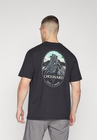 T-shirt en coton noir avec un graphique d'une montagne à l'intérieur d'un ovale, avec le texte "Chouinard" et "Construit pour endurer" en bas.