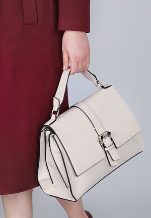 Witte leren handtas met zwarte accenten, gestructureerde vorm, bovenhandvat en een decoratieve gesp. Vastgehouden door een hand tegen een bordeauxkleurige jas.