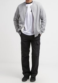 Šedá bomber bunda, bílý tričko a černé cargo kalhoty s bočními kapsami. Černé sneakers doplňují celý outfit. Neformální, funkční styl.