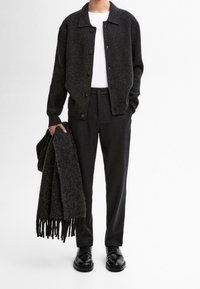 Dark, ribbed cardigan, white t-shirt, tailored dark trousers, holding a textured black scarf with fringes, and wearing black shoes.