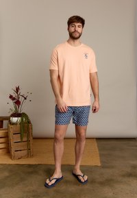 Perzikkleurig t-shirt met een kleine grafische print, gebloemde blauwe zwembroek en blauwe flip-flops. Staand tegen een neutrale achtergrond.