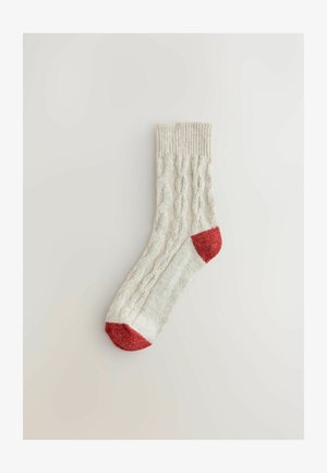 Chaussette en maille torsadée crème avec une bordure côtelée, présentant un orteil et un talon rouges unis. Le matériau texturé ajoute un intérêt visuel et de la chaleur.
