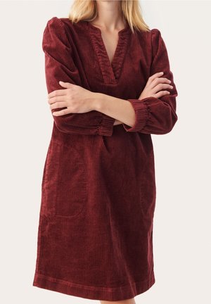 Kobieta ubrana w bordową sukienkę z sztruksu na długim rękawie z dekoltem w serek i dwoma przednimi kieszeniami, ze skrzyżowanymi rękami na piersi.