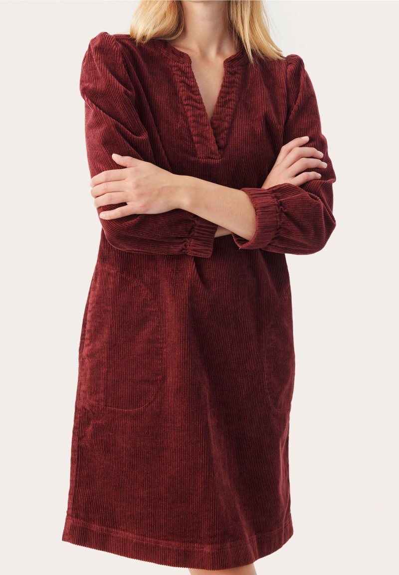 Femme portant une robe en velours côtelé bordeaux à manches longues, col en V et deux poches frontales, bras croisés sur la poitrine.