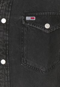 Svart denimjacka med en bröstficka som har en liten broderad logotyp, metallknappar och en något blekt textur.