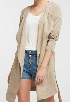 Manteau long beige à ouverture frontale avec col drapé, porté sur un haut blanc et un short en denim bleu taille haute avec boutons sur le devant.