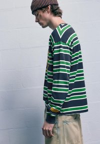 Dlhý rukáv balónového trička prúžkovaného v tmavomodrej, zelenej a bielej farbe, s žltým logom "Stranger Things" na rukáve; nosené s svetlými béžovými šortkami.