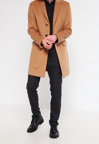 Kamelrock med slag och två svarta knappar, bärs över en svart skjorta; ihop med mörka mönstrade byxor och svarta skor.