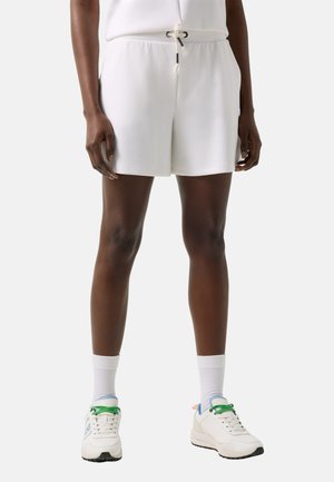 Persoon draagt witte shorts met trekkoord, witte sokken tot aan de kuit en witte sneakers met groene veters, staande tegen een effen achtergrond.