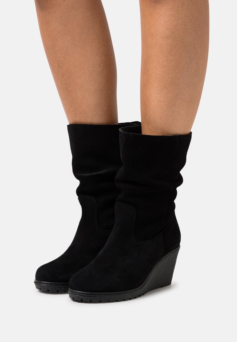 Bottes compensées en daim noir mi-mollet avec bouts ronds portées sur jambes nues sur un fond clair uni.