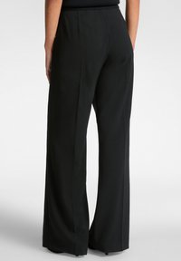 Personne portant un pantalon large noir, vue de dos, se tenant devant un fond clair.