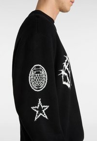 Suéter negro con gráficos bordados en blanco: emblema circular con un patrón de cuadros y un contorno de estrella en las mangas. Material de punto texturizado.
