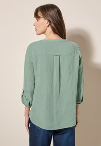 Blusa verde, con texture, maniche a tre quarti, scollatura rotonda e piega centrale sulla schiena. L'orlo è leggermente curvato nella parte inferiore.