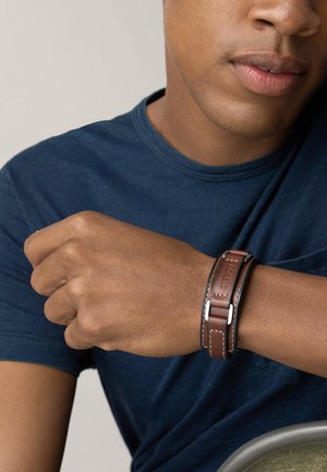 Bracciale in pelle marrone con accenti in metallo argentato; presenta cuciture a contrasto e un design a doppio avvolgimento. Indossato su un polso.