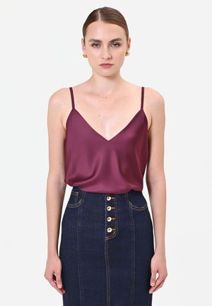 Donna con pelle chiara e capelli tirati indietro indossa orecchini d'oro, un top senza maniche in raso viola scuro e una gonna in denim scuro a vita alta con bottoni.