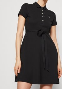 Czarna sukienka polo z krótkim rękawem, z zapięciem na guziki i przewiązywanym paskiem w talii. Wykonana z gładkiego materiału, z dopasowanym górnym partem i rozszerzaną spódnicą.