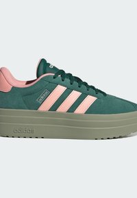 Baskets Adidas vertes et roses avec une semelle épaisse olive, trois bandes roses sur le côté et des lacets noirs sur un fond clair uni.