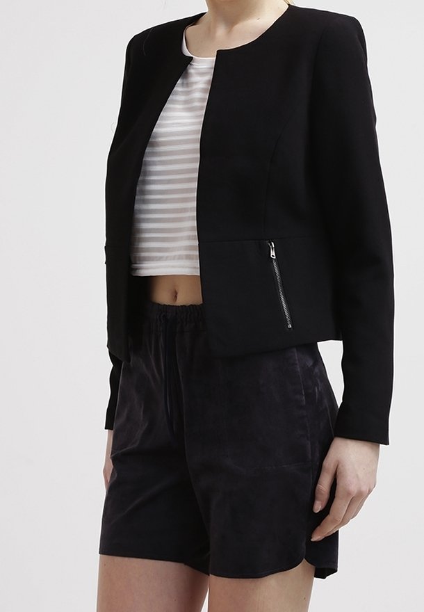 Personne portant une veste noire zippée sur un crop top rayé blanc et gris et un short noir ample devant un fond uni.