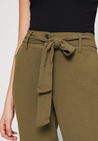 Olijfgroene broek met knoopdetail aan de voorkant bij de taille, textuurstof en een knoopsluiting, gecombineerd met een zwart shirt.