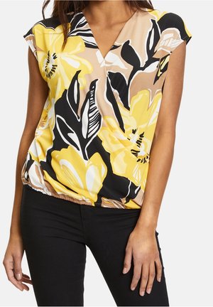 Frau trägt ärmellose Bluse mit V-Ausschnitt und großem gelb-schwarzem Blumenmuster, kombiniert mit schwarzer Hose.