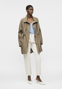 Žena oblečená v béžovom nadrozmernom kabáte, modrom top s čipkovaným lemom, bielych nohaviciach s vysokým pásom a bielych špicatých topánkach na nízkom podpätku.