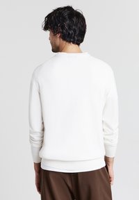 Mann mit dunklem, lockigem Haar, der einen weißen Langarmpullover und braune Hosen trägt und von einer einfarbigen, hellgrauen Hintergrund abgewandt ist.