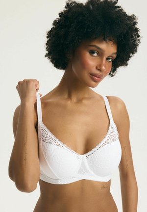 Žena s kučeravými vlasmi nosiaca biele čipkované podprsenku, ktorá si pravou rukou upravuje ramienko na neutrálnom pozadí.