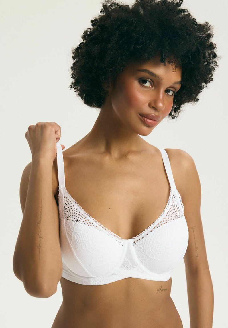 Žena s kučeravými vlasmi nosiaca biele čipkované podprsenku, ktorá si pravou rukou upravuje ramienko na neutrálnom pozadí.